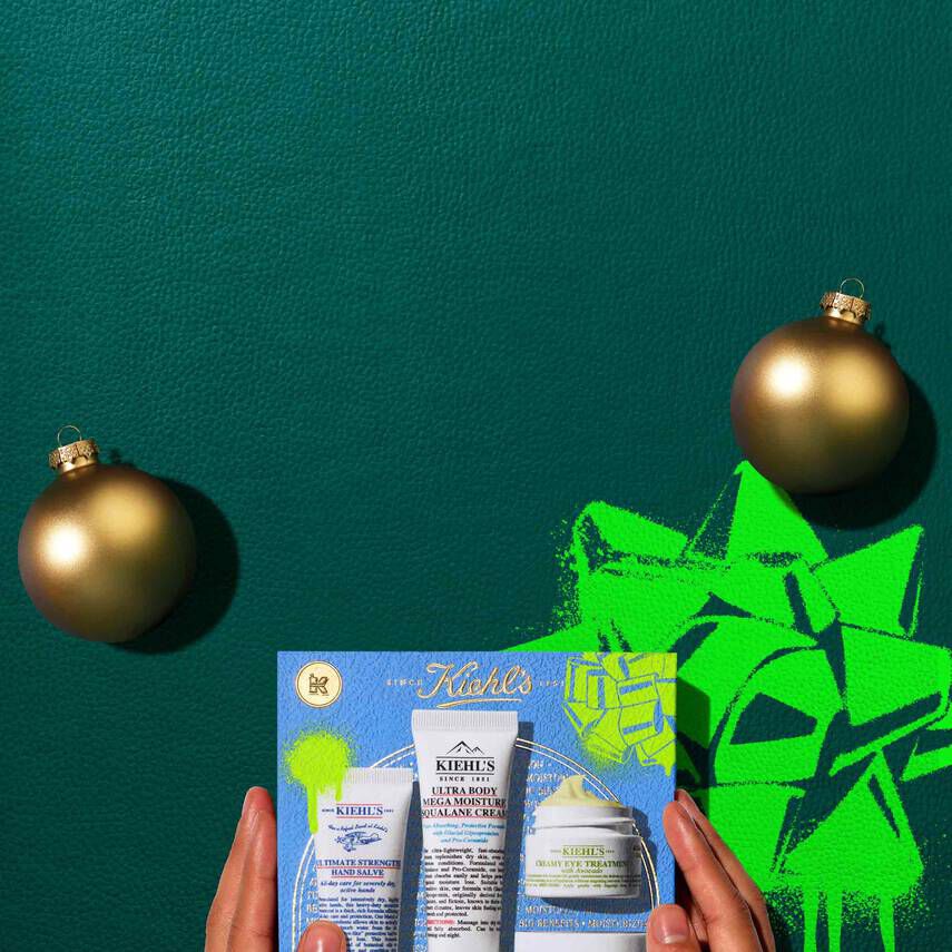 A graphic, set against a dark green textured background, features four Kiehl’s products and a gift box. The top row displays a white tube of "KIEHL’S ULTIMATE STRENGTH HAND SALVE," a white tube of "KIEHL’S ULTRA BODY MEGA MOISTURE SQUALANE CREAM," a small open white jar of "KIEHL’S CREAMY EYE TREATMENT WITH AVOCADO" with visible pale yellow cream, and a white open jar of "KIEHL’S ULTRA FACIAL CREAM" with visible white cream. Two gold spherical ornaments are also present. Below these, a blue Kiehl’s gift box showcases the same four products, decorated with bright green graphic paint splatters and a bow design.