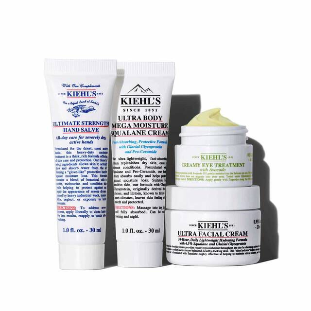 A graphic featuring four white Kiehl’s skincare products arranged on a white background. From left to right: a tube of "KIEHL’S ULTIMATE STRENGTH HAND SALVE," a tube of "KIEHL’S ULTRA BODY MEGA MOISTURE SQUALANE CREAM," a small jar of "KIEHL’S CREAMY EYE TREATMENT WITH AVOCADO" with visible pale yellow cream, and a larger jar of "KIEHL’S ULTRA FACIAL CREAM." The eye cream and facial cream jars are stacked.