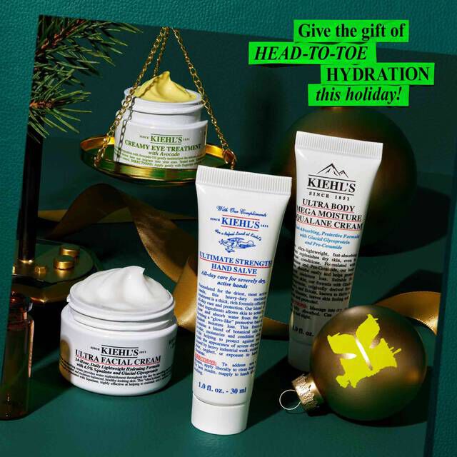 A graphic, set against a dark green textured background, features several Kiehl's products and holiday decorations. A small open white jar of "KIEHL’S CREAMY EYE TREATMENT WITH AVOCADO" with visible pale yellow cream is balanced on a gold scale with a pine branch. A white jar of "KIEHL’S ULTRA FACIAL CREAM" with visible white cream, a white tube of "KIEHL’S ULTIMATE STRENGTH HAND SALVE," and a white tube of "KIEHL’S ULTRA BODY MEGA MOISTURE SQUALANE CREAM" are also arranged on the surface. A gold spherical ornament with a green "K" logo and a dark ribbon are part of the festive display. In the upper right corner, a bright green box contains the text "Give the gift of HEAD-TO-TOE HYDRATION this holiday!"