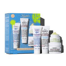 A graphic featuring a blue Kiehl’s gift box green accents on the left. The box contains four products. To the right of the box, the same four products are displayed outside of the box: a tube of "KIEHL’S ULTIMATE STRENGTH HAND SALVE," a tube of "KIEHL’S ULTRA BODY MEGA MOISTURE SQUALANE CREAM," a jar of "KIEHL’S CREAMY EYE TREATMENT WITH AVOCADO," and a jar of "KIEHL’S ULTRA FACIAL CREAM." The box is decorated with bright green graphic paint splatters and a bow design, and features the text "MINI MOISTURES, BIG BENEFITS" at the bottom.