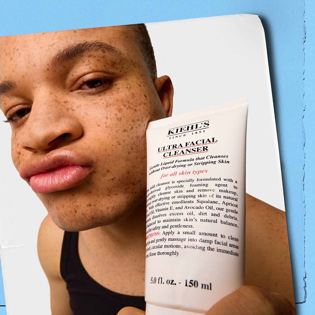 Hand holding a tube of Kiehl’s Ultra Facial Cleanser against a blue and white background.